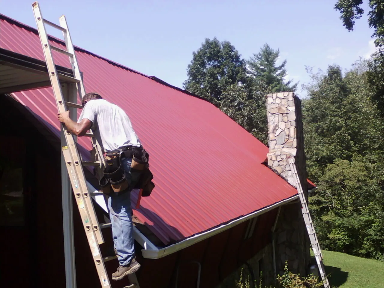 Roofer on a ladder beside a red metal roof and stone chimney — Soffit & Fascia Repair by Roofing Brothers in Koosharem